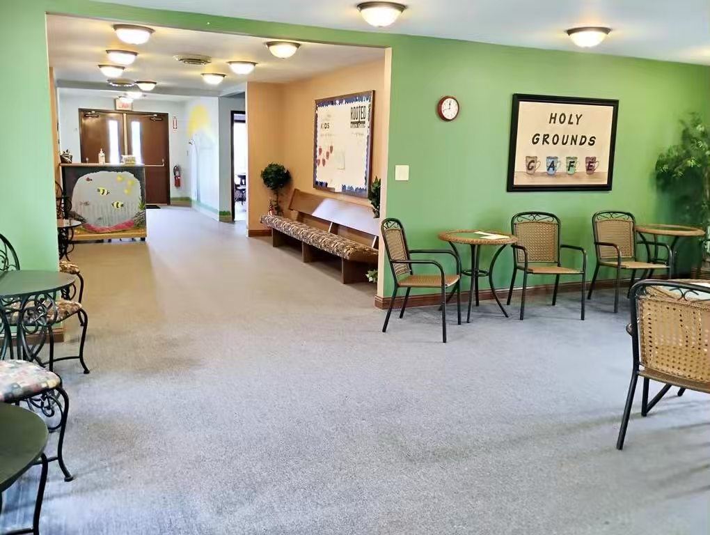 Bright green café seating area with bistro tables and chairs and “Holy Grounds Cafe” sign. Down a hallway, additional seating and a bulletin board are visible.