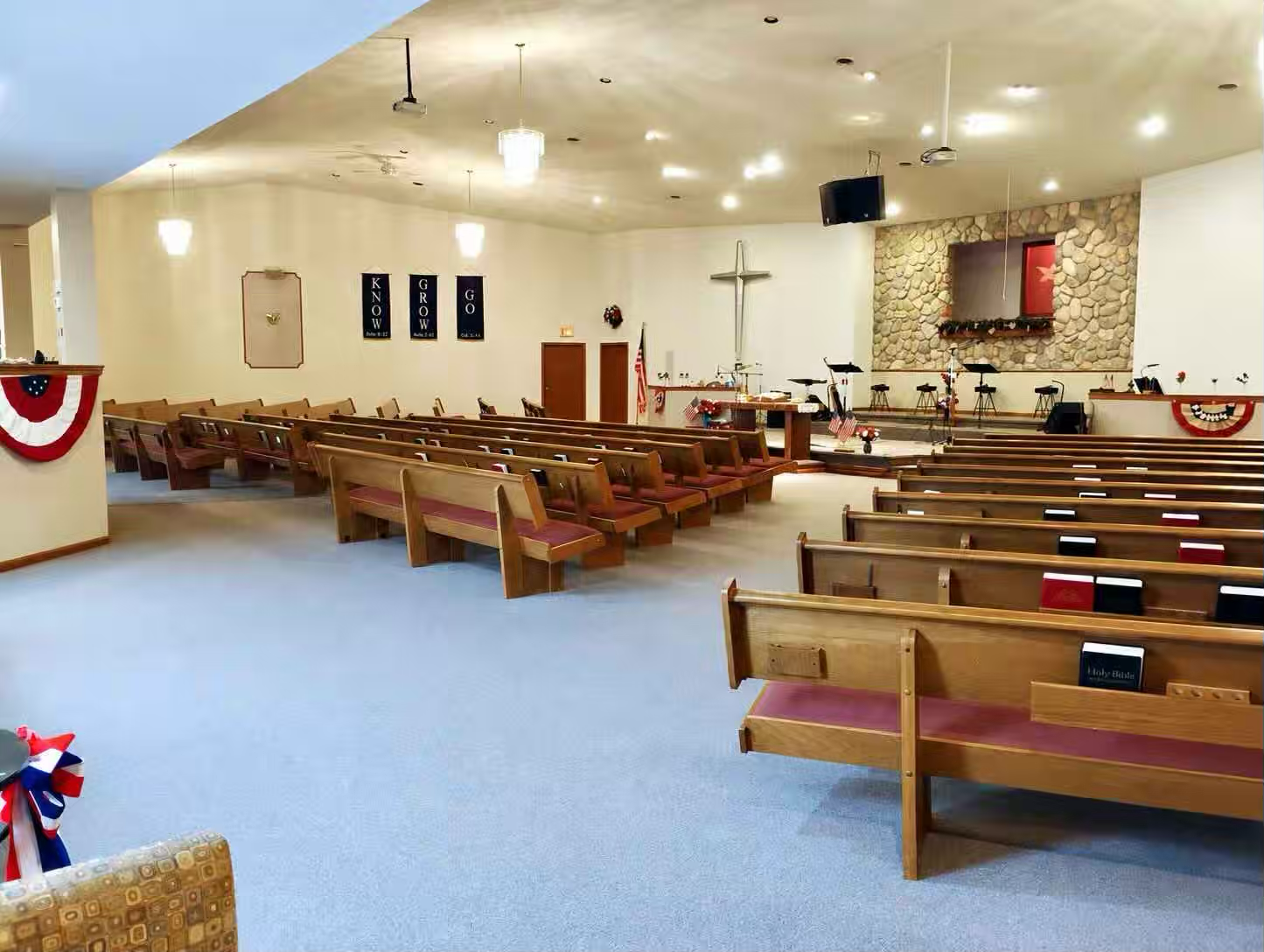 Empty church sanctuary with altar area, baptistry, and wooden pews holding Bibles and song books. A silver cross and blue banners hang on the walls under bright ceiling lights.